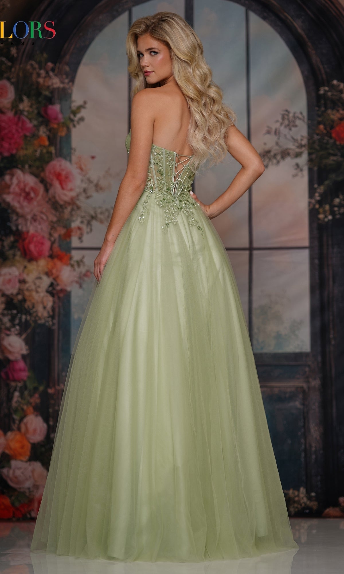 A woman in the Strapless Long Corset Prom Dress: Colors Dress 3923, featuring a lace-up back, stands with her back to the camera before a floral and window backdrop.